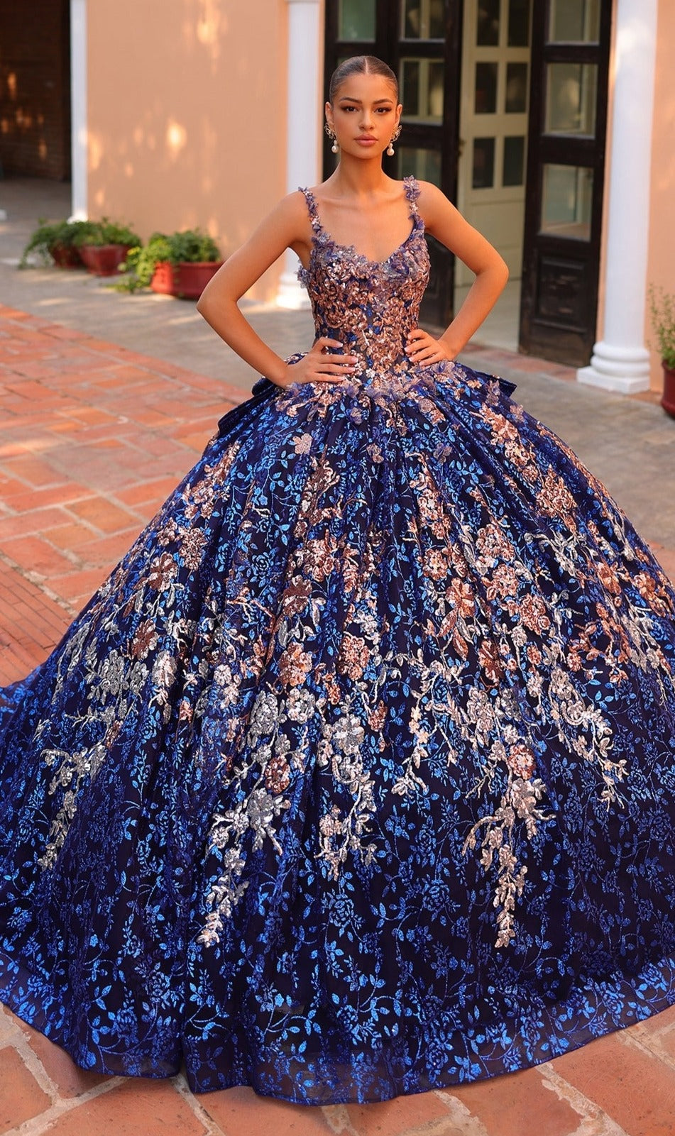 A woman stands outdoors in the Quinceanera Dress 54322 by Amarra, a stunning blue ball gown with floral embroidery and a voluminous, intricately detailed skirt.